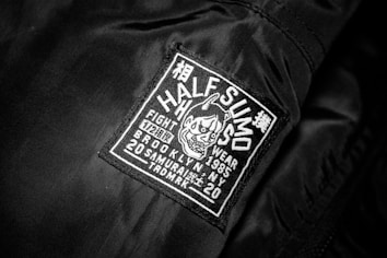 A black jacket features a rectangular patch with white embroidery. The patch includes text and a graphic of an aggressive-looking character with horns. The text on the patch reads 'HALF SUMO', 'FIGHT WEAR', '1985', 'BROOKLYN NY', '20 SAMURAI', and other smaller characters, contributing to a bold and edgy appeal.