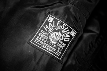A black jacket features a rectangular patch with white embroidery. The patch includes text and a graphic of an aggressive-looking character with horns. The text on the patch reads 'HALF SUMO', 'FIGHT WEAR', '1985', 'BROOKLYN NY', '20 SAMURAI', and other smaller characters, contributing to a bold and edgy appeal.