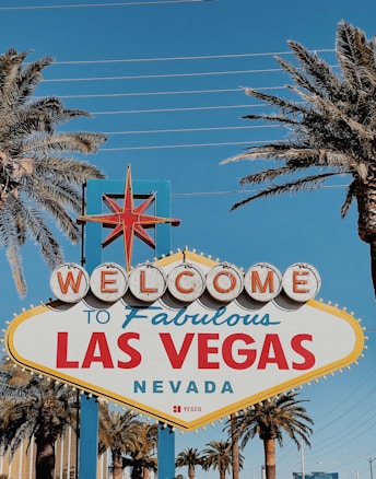 A retro-style welcome sign with the text 'Welcome to Fabulous Las Vegas, Nevada' is surrounded by tall palm trees against a clear blue sky.