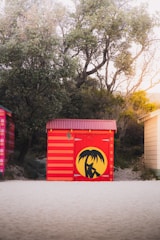 A vibrant red and yellow beach hut stands on a sandy shore, featuring a painted palm tree design on its door. It is surrounded by lush greenery and tall trees, casting soft shadows with the warm light of the setting or rising sun.