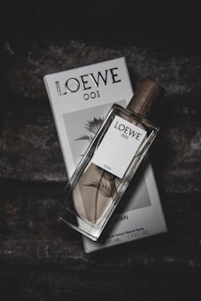 A rectangular glass bottle of perfume with a wooden cap lays diagonally on its corresponding packaging box. The bottle has a white label with text and a botanical illustration. The surrounding background is dark and textured, providing a contrast to the light-colored bottle and box.