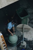 A person is wearing a cap and face covering while lifting the lid of a green trash can. Nearby is another large green bin and a broom leaning against it. The setting appears to be outdoors on a concrete surface with an overhead view, including a tall signpost and a ladder to the side.