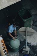 A person is wearing a cap and face covering while lifting the lid of a green trash can. Nearby is another large green bin and a broom leaning against it. The setting appears to be outdoors on a concrete surface with an overhead view, including a tall signpost and a ladder to the side.