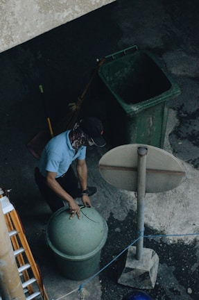 A person is wearing a cap and face covering while lifting the lid of a green trash can. Nearby is another large green bin and a broom leaning against it. The setting appears to be outdoors on a concrete surface with an overhead view, including a tall signpost and a ladder to the side.