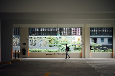A person walks across an exposed car park area with a modern, urban residential backdrop. The area is well-lit with natural light from the large windows. There are plants visible in the background through the openings.