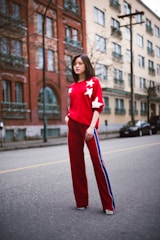 A person stands confidently in the middle of an urban street. They are wearing a stylish red outfit consisting of a sweater with white stars and matching pants with blue and white stripes along the sides. The background features a mix of red brick and beige buildings with several windows and balconies.