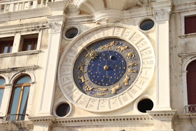 An ornate astronomical clock with a deep blue and gold design featuring zodiac symbols. The clock is set in a historical building facade with Roman numerals and intricate architectural details.