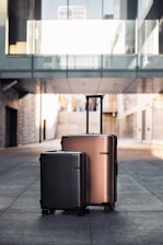 Two suitcases are placed side by side on a paved walkway in an urban setting. The larger suitcase is metallic brown and the smaller one is black. The background features modern architectural elements with a glass bridge and buildings.