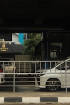 A street scene features a stationary white vehicle partially visible behind a metal railing. Nearby, another vehicle is parked under an overpass. Behind the railing, a blue and black building facade displays an electrical power sign. The surrounding environment includes some greenery and hints of urban infrastructure.