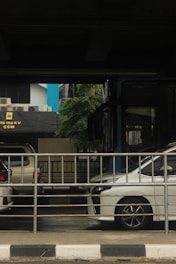 A street scene features a stationary white vehicle partially visible behind a metal railing. Nearby, another vehicle is parked under an overpass. Behind the railing, a blue and black building facade displays an electrical power sign. The surrounding environment includes some greenery and hints of urban infrastructure.