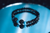 A pair of black and blue beaded bracelets is placed on a reflective blue surface. The beads are shiny and spherical, with larger blue beads interspersed among smaller black ones, creating a visually striking contrast.