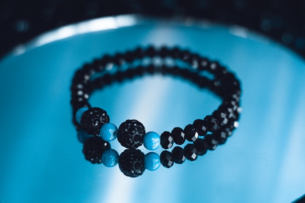 A pair of black and blue beaded bracelets is placed on a reflective blue surface. The beads are shiny and spherical, with larger blue beads interspersed among smaller black ones, creating a visually striking contrast.