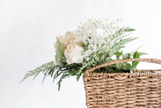 A wicker basket is filled with delicate white roses, baby's breath, and green ferns. The arrangement is simple yet elegant, emphasizing a fresh and natural aesthetic against a plain white background.