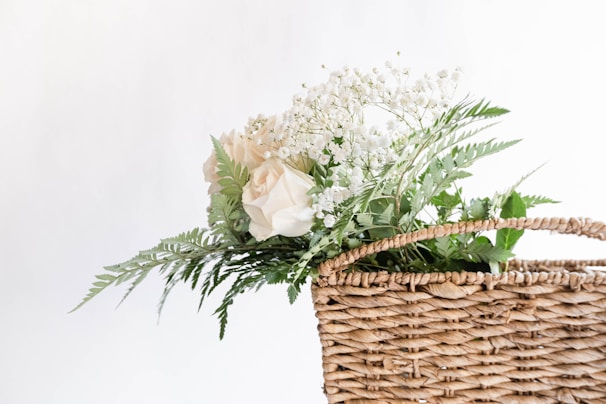 A wicker basket is filled with delicate white roses, baby's breath, and green ferns. The arrangement is simple yet elegant, emphasizing a fresh and natural aesthetic against a plain white background.