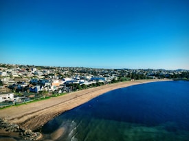 The image features a coastal town with a sandy beach running along the shoreline. Several residential and commercial buildings are visible in the background, indicating a densely populated area. The clear blue sky and vivid turquoise water create a serene atmosphere. A few small waves can be seen lapping onto the shore, with some rock formations at the edge of the beach.