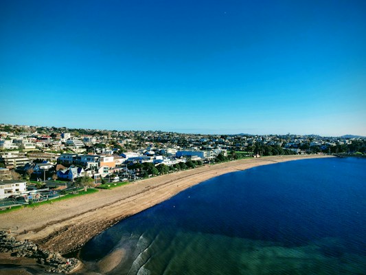 The image features a coastal town with a sandy beach running along the shoreline. Several residential and commercial buildings are visible in the background, indicating a densely populated area. The clear blue sky and vivid turquoise water create a serene atmosphere. A few small waves can be seen lapping onto the shore, with some rock formations at the edge of the beach.