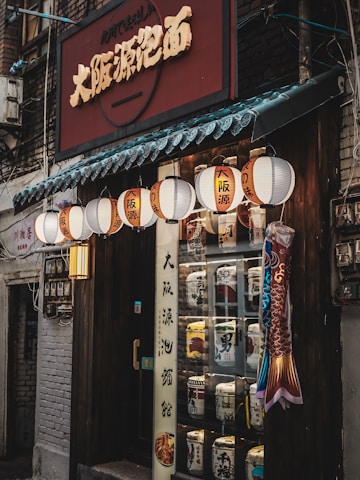 An inviting storefront filled with traditional Japanese lanterns adorned with kanji characters, a sign bearing bold lettering, and a display of decorative barrels. The wooden exterior exudes a rustic charm, and a colorful fish decoration adds a festive touch.