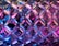 A close-up view of a textured surface resembling a glass or plastic material with a repeating diamond pattern. The surface is illuminated with vibrant neon colors, including pink, blue, and purple, creating a luminous and dynamic effect.