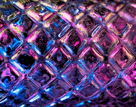 A close-up view of a textured surface resembling a glass or plastic material with a repeating diamond pattern. The surface is illuminated with vibrant neon colors, including pink, blue, and purple, creating a luminous and dynamic effect.