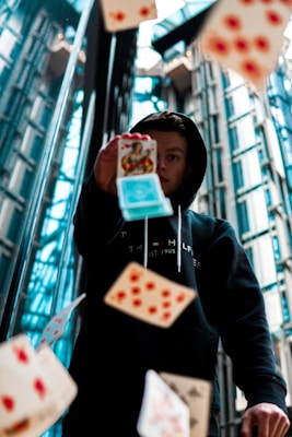 A person in a dark hoodie is surrounded by playing cards flying through the air in an urban environment. The glass, steel, and modern architecture reflect an industrial, futuristic setting.