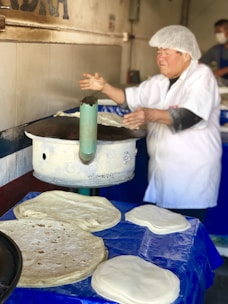A person wearing a white hat and uniform is making tortillas on a large, round, metal cooking surface. Several uncooked tortillas are placed on a blue tablecloth in front of the person. The background shows a tiled wall with part of a word visible.
