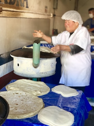 A person wearing a white hat and uniform is making tortillas on a large, round, metal cooking surface. Several uncooked tortillas are placed on a blue tablecloth in front of the person. The background shows a tiled wall with part of a word visible.