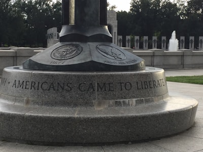 A monument with engravings featuring emblems of the Navy and Army Air Forces, including an inscription that reads 'AMERICANS CAME TO LIBERATE'. The setting appears to be an outdoor memorial area with stone structures and a visible fountain in the background.