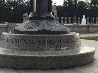 A monument with engravings featuring emblems of the Navy and Army Air Forces, including an inscription that reads 'AMERICANS CAME TO LIBERATE'. The setting appears to be an outdoor memorial area with stone structures and a visible fountain in the background.