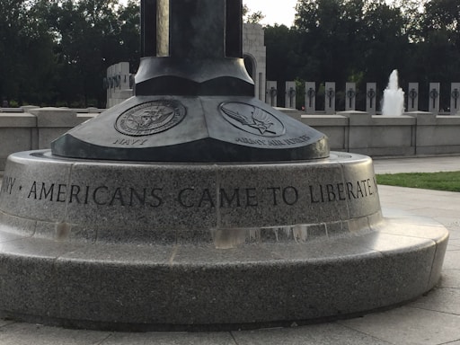 A monument with engravings featuring emblems of the Navy and Army Air Forces, including an inscription that reads 'AMERICANS CAME TO LIBERATE'. The setting appears to be an outdoor memorial area with stone structures and a visible fountain in the background.
