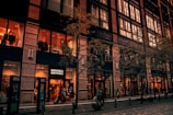 A multi-story building with large storefront windows showcasing interior lighting and clothing displays. The sign 'Brooks Brothers' indicates a store entrance, with people walking along the sidewalk. Trees with sparse foliage line the street in front of the building.