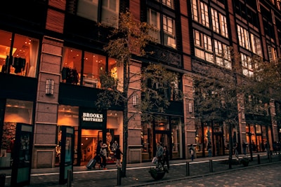 A multi-story building with large storefront windows showcasing interior lighting and clothing displays. The sign 'Brooks Brothers' indicates a store entrance, with people walking along the sidewalk. Trees with sparse foliage line the street in front of the building.
