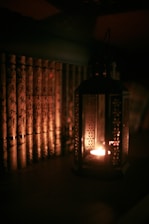 A dimly lit scene featuring a row of books with ornate covers arranged vertically side by side. To the right of the books, a decorative lantern emits a warm, soft glow from a small candle inside, casting subtle shadows and contributing to an intimate ambiance.