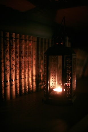 A dimly lit scene featuring a row of books with ornate covers arranged vertically side by side. To the right of the books, a decorative lantern emits a warm, soft glow from a small candle inside, casting subtle shadows and contributing to an intimate ambiance.
