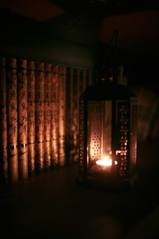 A dimly lit scene featuring a row of books with ornate covers arranged vertically side by side. To the right of the books, a decorative lantern emits a warm, soft glow from a small candle inside, casting subtle shadows and contributing to an intimate ambiance.