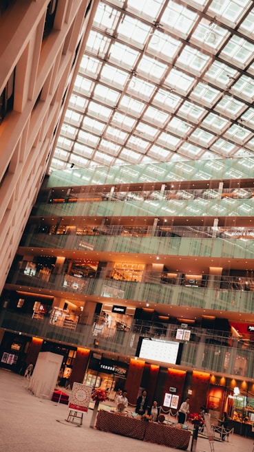 A spacious multi-level shopping mall interior with a high glass ceiling allowing natural light to enter. The floors are lined with various shops and boutiques. The ground level features a setup with a table decorated for an event, with people engaged in an activity.