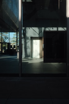 The image captures a minimalistic architectural scene with strong shadows and reflections on a glass facade. The lighting creates an interplay of light and shadow, highlighting the clean, geometric lines of the structure. A portion of the floor is illuminated by sunlight, while the rest of the space is shrouded in darkness, creating a high contrast effect.