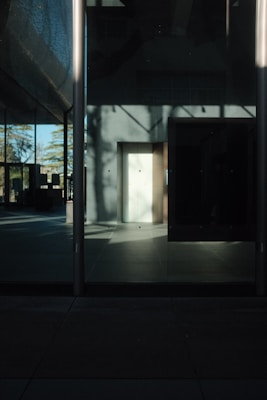 The image captures a minimalistic architectural scene with strong shadows and reflections on a glass facade. The lighting creates an interplay of light and shadow, highlighting the clean, geometric lines of the structure. A portion of the floor is illuminated by sunlight, while the rest of the space is shrouded in darkness, creating a high contrast effect.