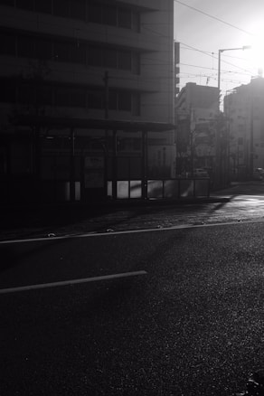 A quiet urban street scene captured during early morning or late afternoon. The sun casts long shadows across the road, and the buildings in the background are partially illuminated. The overall atmosphere is calm and serene, with a mostly empty street.