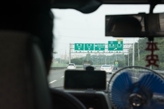 View from inside a vehicle driving on a highway with green directional road signs ahead, indicating locations such as Ligang, Yancha, and Dashe. The image shows a number of cars traveling in both lanes. In the foreground, part of the vehicle's interior is visible, including a rearview mirror, a small fan, and a hanging ornament.