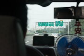 View from inside a vehicle driving on a highway with green directional road signs ahead, indicating locations such as Ligang, Yancha, and Dashe. The image shows a number of cars traveling in both lanes. In the foreground, part of the vehicle's interior is visible, including a rearview mirror, a small fan, and a hanging ornament.