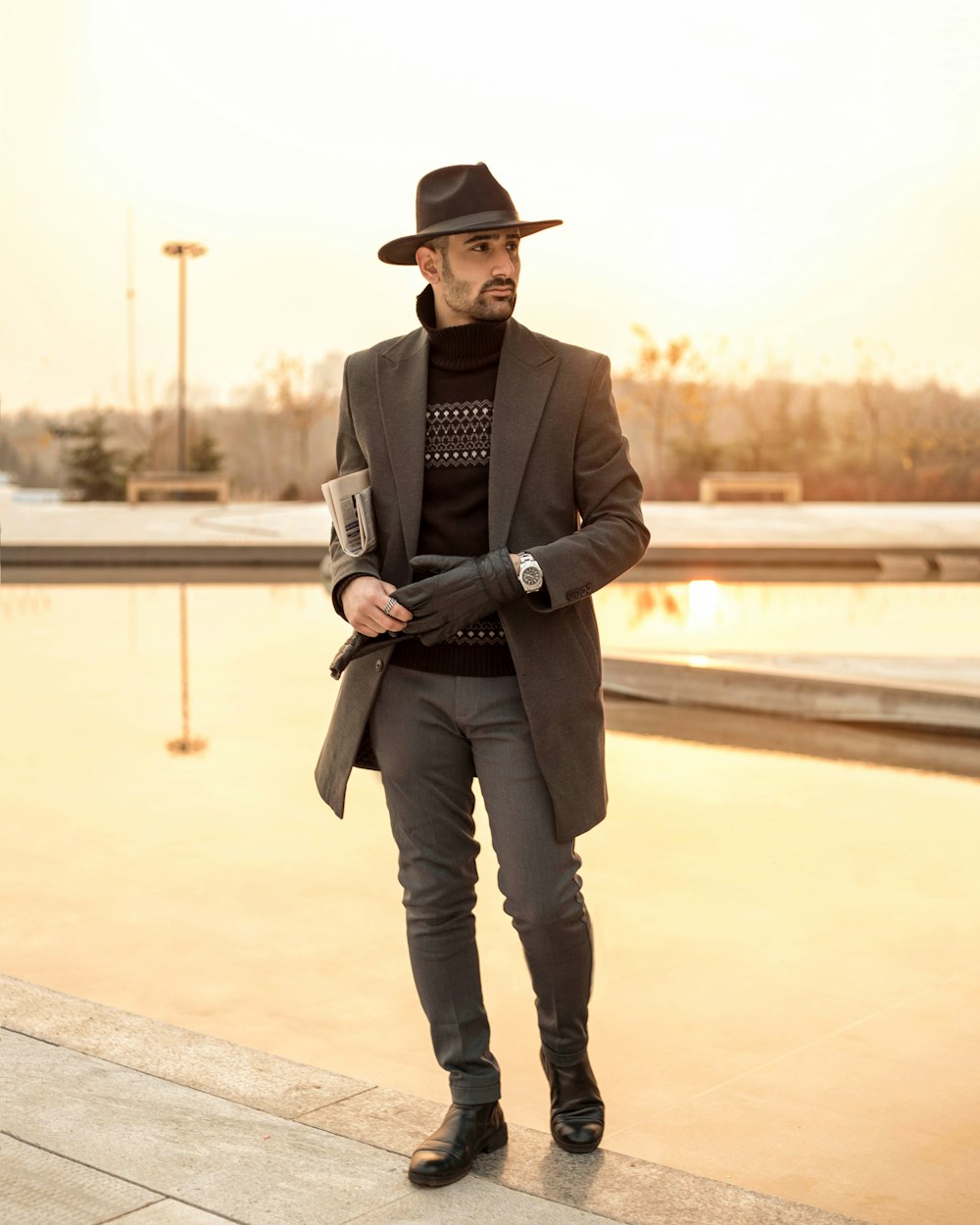 Homme en blazer noir et pantalon gris portant un chapeau fedora noir debout sur un trottoir en béton gris
