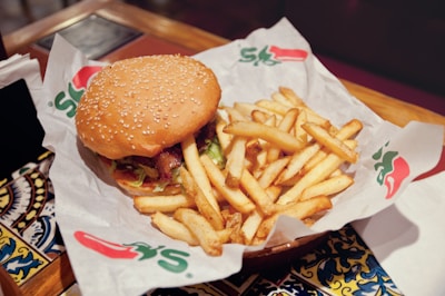 A sesame seed hamburger bun is filled with lettuce, tomato, cheese, bacon, and a beef patty, placed beside a generous serving of golden french fries. The food is presented on branded paper in a restaurant setting, with decorative tiles visible underneath the serving tray.