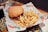 A sesame seed hamburger bun is filled with lettuce, tomato, cheese, bacon, and a beef patty, placed beside a generous serving of golden french fries. The food is presented on branded paper in a restaurant setting, with decorative tiles visible underneath the serving tray.