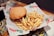 A sesame seed hamburger bun is filled with lettuce, tomato, cheese, bacon, and a beef patty, placed beside a generous serving of golden french fries. The food is presented on branded paper in a restaurant setting, with decorative tiles visible underneath the serving tray.