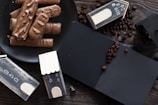 A black plate filled with chocolate-coated wafer bars is placed on a wooden surface. Surrounding the plate are miniature black wooden houses with white roof accents. Coffee beans are scattered around, adding a rustic touch to the arrangement. A black notebook partially covered in coffee beans is also present.