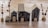 A collection of small, handcrafted wooden houses is arranged on a wooden surface. The houses vary in size, have dark walls with light-colored roofs, and feature painted windows and doors. Surrounding the houses are small decorative trees with brown branches and round bases.