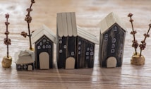 A collection of small, handcrafted wooden houses is arranged on a wooden surface. The houses vary in size, have dark walls with light-colored roofs, and feature painted windows and doors. Surrounding the houses are small decorative trees with brown branches and round bases.