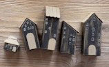 A series of five small, handcrafted wooden houses with black bodies and beige roofs, placed on a wooden surface. Each house is decorated with simple white window and door designs, giving a quaint village-like appearance.