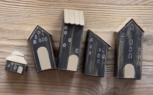 A series of five small, handcrafted wooden houses with black bodies and beige roofs, placed on a wooden surface. Each house is decorated with simple white window and door designs, giving a quaint village-like appearance.