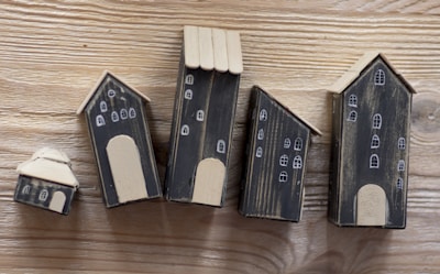 A series of five small, handcrafted wooden houses with black bodies and beige roofs, placed on a wooden surface. Each house is decorated with simple white window and door designs, giving a quaint village-like appearance.